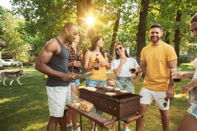Group of happy friends having beer and barbecue party with party rentals set up at sunny day. Resting together outdoor in a forest glade or backyard. Celebrating and relaxing, laughting. Summer lifestyle, friendship concept.